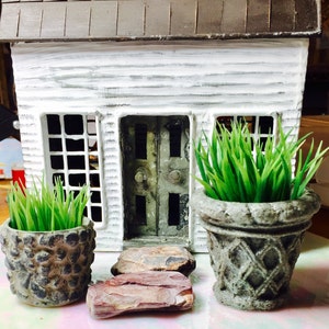 May include: A miniature white house with a brown roof and a weathered appearance. Two small pots with green grass flank the house, and a set of stone steps lead to the front door. The house has a rustic, handcrafted aesthetic.