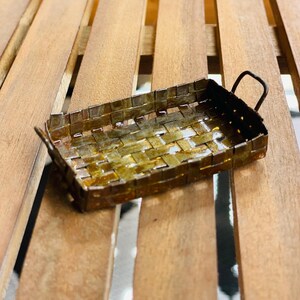 May include: A small, rectangular metal basket with a woven design. The basket has a dark, aged appearance with a handle on each end. The basket is sitting on a wooden surface.