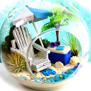 May include: A miniature beach scene inside a glass terrarium. It features a white Adirondack chair, a blue and white umbrella, a cooler, sunglasses, and a palm tree. The base is sand with blue glass accents, seashells, and flip-flops.
