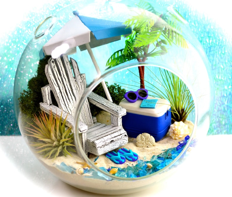May include: A miniature beach scene inside a glass terrarium. It features a white Adirondack chair, a blue and white umbrella, a cooler, sunglasses, and a palm tree. The base is sand with blue glass accents, seashells, and flip-flops.