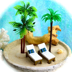 May include: Miniature beach scene with two white lounge chairs with blue and green striped pillows, two palm trees, and a seashell on a sandy surface. A piece of driftwood and a coral-like object are also present.