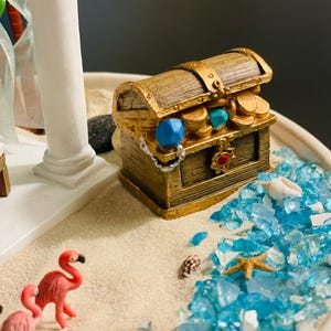 May include: A miniature treasure chest with gold accents, filled with coins and jewels, sits on a sandy beach scene. Pink flamingos, blue glass, seashells, and a starfish add to the coastal theme. A white column is in the background.
