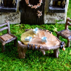 May include: Miniature fairy garden scene with a leaf-shaped table and two twig chairs. The table holds three tiny blue teacups. The scene is set on a bed of green moss, with a small house in the background.