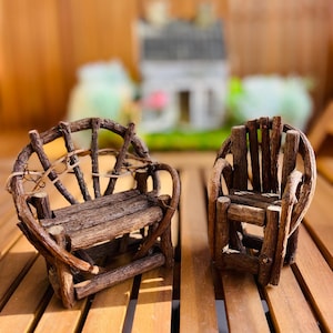 May include: Two miniature wooden chairs, one a bench and the other an armchair, are displayed on a wooden surface. The chairs are crafted from small branches and twigs, giving them a rustic appearance. A miniature house is visible in the background.