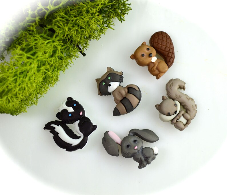May include: A collection of six woodland animal-themed embellishments. The set includes a skunk, raccoon, squirrel, beaver, and a rabbit. The animals are in various shades of brown, gray, black, and white, with some pink accents. The embellishments are on a white surface.