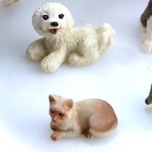 May include: Miniature figurines of a white dog and a tan cat. The dog is lying down with its mouth open, and the cat is in a resting pose. The figurines are detailed and appear to be made of a resin material.