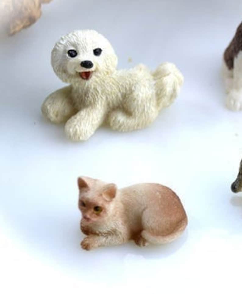 May include: Miniature figurines of a white dog and a tan cat. The dog is lying down with its mouth open, and the cat is in a resting pose. The figurines are detailed and appear to be made of a resin material.