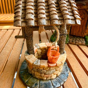 May include: A miniature wishing well with a stone-like appearance. The well features a weathered, tiled roof, wooden supports, and a small orange bucket hanging from a rope. The base is a circular stone platform.