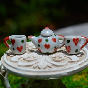 May include: A miniature white ceramic tea set with red heart and green dot designs. The set includes a teapot and two teacups, arranged on a small, ornate, white table. The teacups have red handles. The background is blurred green.