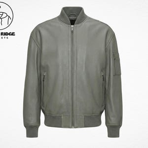 Peut inclure: Veste de bombardier en cuir gris avec fermeture éclair, deux poches latérales et col et poignets côtelés. La veste a un design classique et présente le logo "Retro Ridge Jackets" dans le coin supérieur gauche.