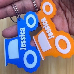 Può includere: Due portachiavi a forma di trattore, uno blu e uno arancione, ciascuno con il nome "Jessica" in bianco. I portachiavi hanno dettagli circolari bianchi e un portachiavi argentato.