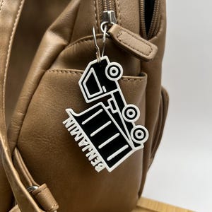 Può includere: Un portachiavi a forma di camion nero e bianco con il nome "BENJAMIN" stampato sopra, appeso a una borsa in pelle marrone. Il portachiavi è attaccato con un fermaglio in metallo color argento.