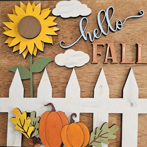 May include: A wooden wall sign featuring a yellow sunflower, two white clouds, and the text "hello FALL" in blue and orange. A white picket fence sits at the bottom with two orange pumpkins, a yellow leaf, a green leaf, and a small brown acorn.