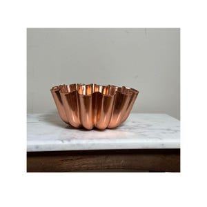 Puede incluir: Un cuenco acanalado de color cobre con un borde festoneado. El cuenco es de metal brillante y se encuentra sobre una superficie de mármol blanco. El diseño del cuenco recuerda a una flor o a un molde decorativo para hornear.