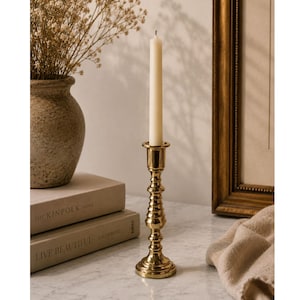 Può includere: Un candelabro dorato con una candela color crema. Il candelabro ha un design dettagliato e ornato. Un vaso con fiori secchi e due libri sono sullo sfondo. Sono visibili anche una cornice e un panno beige.