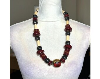 May include: A chunky necklace with a variety of beads in cream, black, and burgundy. The necklace is displayed on a white mannequin. The beads are various shapes, including round, disc, and cylindrical. The focal point is a large, red and gold bead.