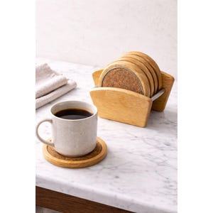 May include: A set of six round coasters with a cork center and a wooden frame, displayed in a wooden holder. A mug of coffee sits on a matching coaster. The coasters are made of wood and cork.