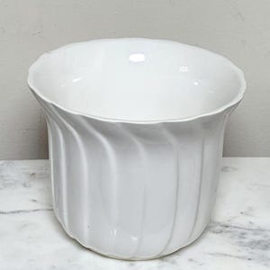Puede incluir: Una taza de cerámica blanca con un asa negra y el texto "But First, Coffee" en negrita negra. La taza está sobre un fondo borroso. La taza mide aproximadamente 10 cm de altura.