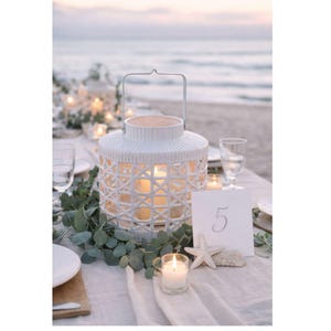 May include: A white lantern with a woven design and a metal handle sits on a table set for an outdoor event. The table is decorated with greenery, candles, and a table number. The background shows the ocean and a soft, diffused light.