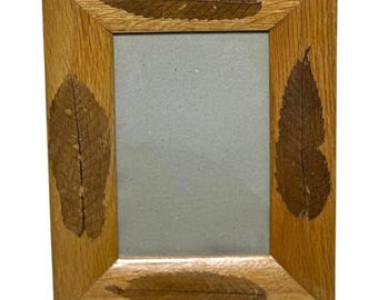 Könnte beinhalten: Ein rechteckiger Bilderrahmen aus hellbraunem Holz mit sichtbarer Holzmaserung. Der Rahmen ist mit vier dunkelbraunen, getrockneten und skelettierten Blättern verziert, die oben, unten, links und rechts in das Holz eingelassen sind.