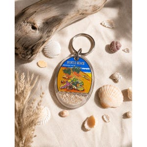 May include: An oval-shaped keychain with a beach scene, the text "MYRTLE BEACH", and sand at the bottom. The keychain is silver and is surrounded by seashells and driftwood on a beige surface.