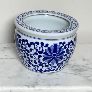 Könnte beinhalten: Ein runder Keramikübertopf mit weißem Untergrund, verziert mit einem tiefblauen Blumenmuster und geschwungenen Rankenmotiven. Der Rand ist mit einem durchgehenden blauen Wirbelmuster versehen, und das Innere ist glatt weiß glasiert.
