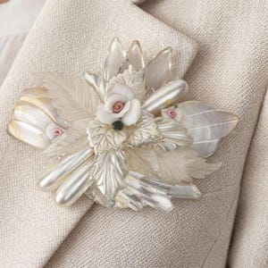 May include: A floral brooch with a pearlescent finish, featuring white roses with pink centers, clear leaves, and a green accent. The brooch is attached to a beige blazer, showcasing its intricate design and craftsmanship.