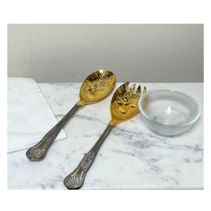 May include: A gold and silver serving spoon and fork set with a clear glass bowl. The spoon and fork have ornate handles and gold-colored serving heads with a grape design. The bowl is small and round.