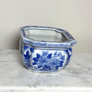 Può includere: Un vaso in ceramica bianca con un motivo floreale e di farfalle blu. Il contenitore esagonale presenta un bordo geometrico a greca lungo il bordo superiore. L'esterno mostra dettagliate illustrazioni blu di fiori in fiore e farfalle.