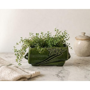 May include: A rectangular, dark green ceramic planter filled with green herbs. The planter has a floral design on the front and small handles on the sides. A small, beige ceramic jar with a lid is in the background.