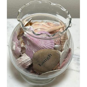 Könnte beinhalten: Eine klare Glasschale mit gerüschtem Rand, gefüllt mit Muscheln in Rosa-, Beige- und Weißtönen. Ein glatter, brauner Stein mit dem eingravierten Wort "Laugh" liegt zwischen den Muscheln.