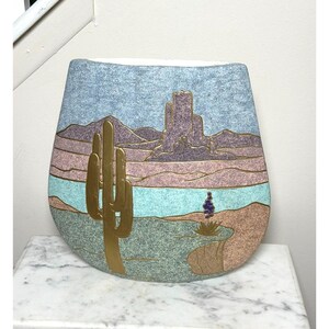 May include: A flat, rounded ceramic vase featuring a desert landscape design. The surface has a textured, speckled finish in shades of blue, purple, and tan. A large, smooth, gold-colored saguaro cactus is embossed on the front.