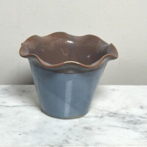 May include: A ceramic bowl featuring a flared, ruffled rim with a glossy brown finish on the interior and upper edge, transitioning into a smooth, muted blue glaze on the exterior body. The vessel has a tapered shape and a wide, open top opening.