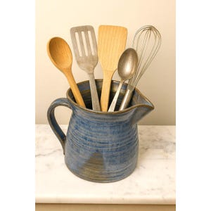 May include: A blue ceramic pitcher holds various kitchen utensils. A wooden spoon, slotted spatula, solid spatula, metal spoon, and whisk are visible. The pitcher has a handle and a spout, and sits on a white surface.