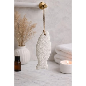 May include: A white, fish-shaped pumice stone hanging from a natural fiber cord. The textured stone is suspended from a brass hook, suggesting a bathroom or spa setting. A candle and towels are in the background.