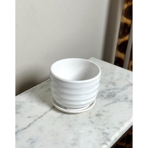 May include: A white ceramic plant pot with a glossy finish and horizontal ribbed texture. The cylindrical container features a matching circular saucer base, providing a clean and simple design for holding small indoor plants or succulents.