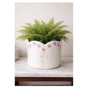 May include: A white ceramic planter with a scalloped rim, adorned with pink and blue floral designs. The planter is filled with lush green ferns. The floral pattern extends down the sides with delicate stem-like details.