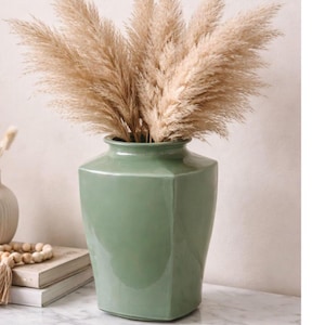 May include: A sage green ceramic vase with a hexagonal shape and a smooth, glossy finish. The vase is filled with a large, dense arrangement of tall, fluffy, beige pampas grass plumes that fan out from the narrow opening at the top of the vessel.