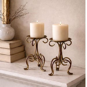 Könnte beinhalten: Zwei Messing-Kerzenhalter mit elfenbeinfarbenen Stumpenkerzen. Die Halter haben ein dekoratives, geschwungenes Design und stehen auf einer weißen Oberfläche. Im Hintergrund ist eine Vase mit getrockneten Zweigen zu sehen.