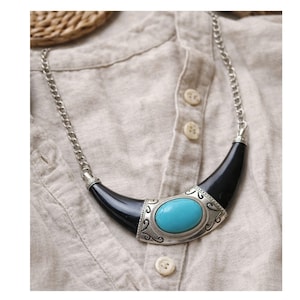 May include: A statement necklace featuring a large oval turquoise stone set in a silver-toned metal frame. The frame is flanked by curved black elements and connected to a silver chain. The necklace rests on a beige fabric.