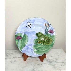 May include: A round ceramic decorative plate featuring a hand-painted scene of a green frog sitting on a large lily pad. The background is a light blue wash with pink water lilies, brown cattails, and a black outlined dragonfly in flight.