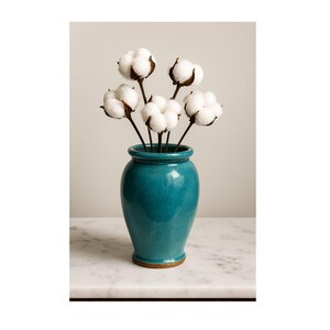 May include: A teal ceramic vase holds a bouquet of cotton stems. The vase has a glossy finish and a rounded shape with a narrow base. The cotton bolls are fluffy and white, creating a contrast with the vase's color.