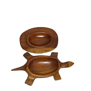 以下が含まれることがあります： 木製のカメ型2ピース食器セット。下の部分は大きな楕円形のボウルが付いたカメです。上の部分は、お揃いの楕円形のくぼみが付いた蓋です。どちらのパーツも濃い茶色の木で作られています。