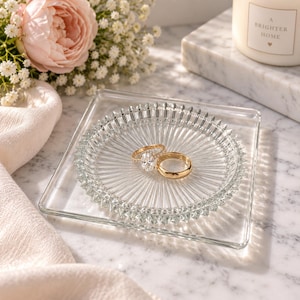 May include: A square clear glass trinket dish with a circular, sunburst-patterned center holds two gold rings, one featuring a central stone. The dish rests on a white marble surface next to a soft fabric and a candle labeled A Brighter Home.