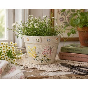 Pode incluir: Vaso de flores de cerâmica creme com ilustrações de borboletas amarelas e rosas. O vaso é preenchido com pequenas flores brancas e está sobre uma toalha de renda. Decorativo e funcional.