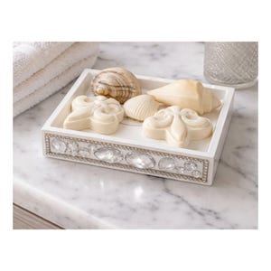 May include: A white decorative tray with a jeweled border holds seashell-shaped soaps and real seashells. The tray is on a marble surface, with a stack of white towels in the background. The soaps are a light cream color.