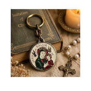 May include: A circular metal keychain pendant featuring a religious image of a woman and child, with the words "Salem" and "Jeru" around the edge. The pendant is attached to a gold-coloured ring and chain. A book, candle, and rosary are in the background.