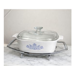 May include: A white casserole dish with a clear glass lid and a blue floral design. The dish sits on a chrome-plated metal stand. The casserole dish is a square shape and has handles on either side.
