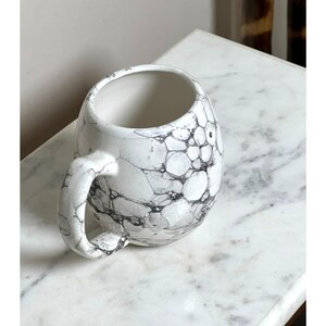 May include: A round ceramic mug featuring a white base with a black bubble or marble-like pattern across the exterior surface. The mug has a curved handle on the side and a wide, open top, showcasing a smooth, glossy finish throughout the piece.