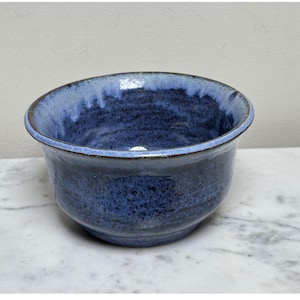 May include: A round ceramic bowl featuring a deep blue speckled glaze with a lighter, pale blue drip pattern along the interior and exterior rim. The bowl has a smooth, curved silhouette and a dark, defined edge around the top opening.